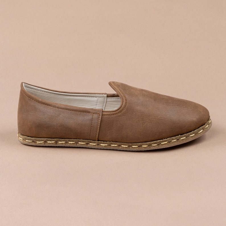 Puede incluir: Un par de zapatos marrones de cuero sin cordones con un detalle de costura blanca alrededor de la suela.
