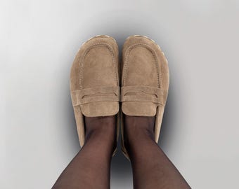 May include: A top-down view of two tan suede loafers. The shoes have a classic design with a rounded toe and a strap detail across the top. The shoes are worn with black sheer hosiery.