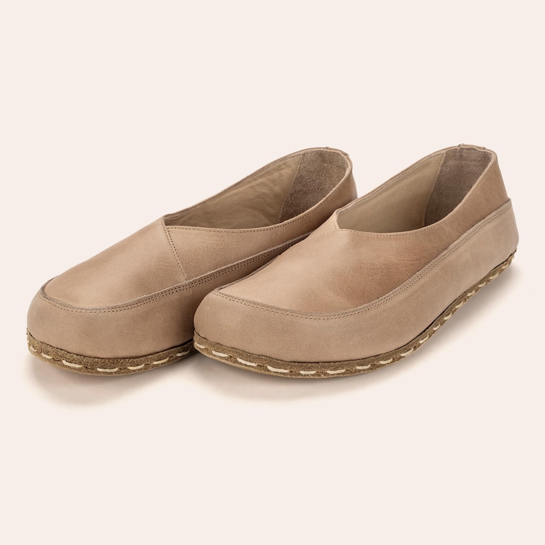 May include: A pair of beige leather slip-on shoes. The shoes have a rounded toe and a stitched sole. The upper part of the shoe is a lighter shade of beige, and the sole is a darker beige with white stitching.
