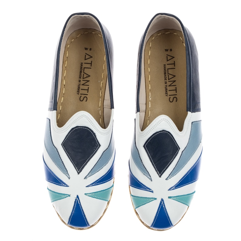 May include: A pair of blue and white leather slip-on shoes with a geometric pattern. The shoes have a white sole and the text "ATLANTIS HANDMADE IN TURKEY" is printed on the inside of each shoe.