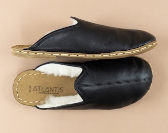 Può includere: Un paio di pantofole in pelle nera con suola in camoscio marrone e fodera in pelle di pecora bianca. Le pantofole hanno un'etichetta che recita "ATLANTIS HANDMADE IN TURKEY".