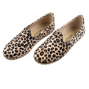 May include: A pair of brown and black leopard print slip-on shoes with a white leather sole. The shoes are made by ATLANTIS and are handmade in Turkey.