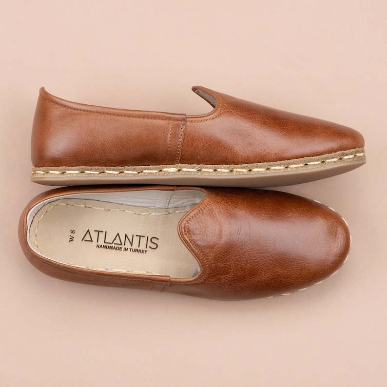May include: A pair of brown leather slippers with white stitching detail around the sole. The slippers are handmade in Turkey and have the brand name "ATLANTIS" printed on the insole.