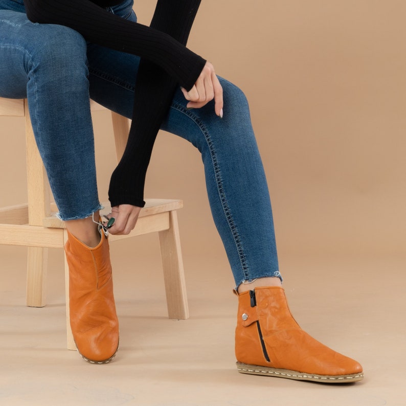 May include: A pair of ankle boots with a zipper on the side. The boots are made of brown leather and have a black sole. The boots are being worn by a person wearing blue jeans.