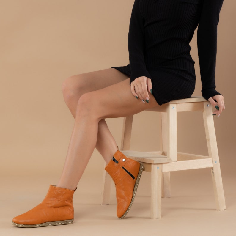 May include: A woman wearing a black dress and orange leather ankle boots sits on a wooden step stool. The boots have a zipper on the side and a small buckle detail.