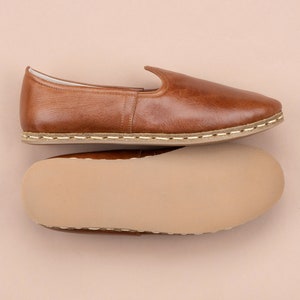 May include: A pair of brown leather slippers with a white stitching detail around the sole. The slippers are shown from the top and bottom, revealing the insole.