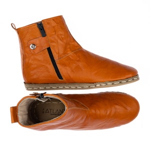 May include: A pair of tan leather ankle boots with a zipper on the side and a stitched seam around the sole. The boots have a label on the inside that says "ATLAN" and "HANDMADE IN".