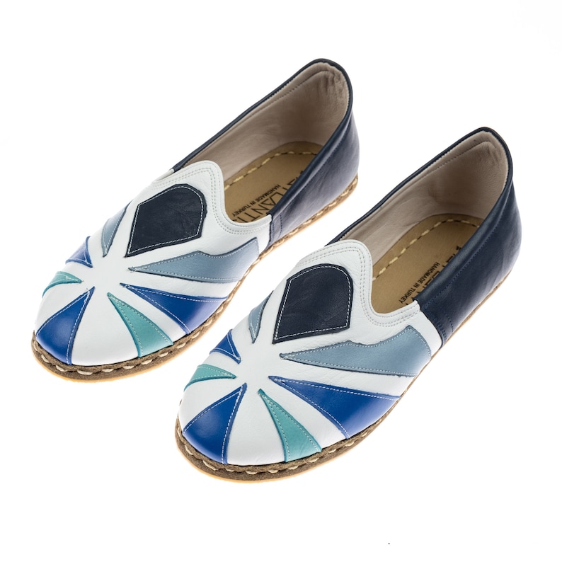 May include: A pair of white leather slip-on shoes with a blue, teal, and gray geometric pattern. The shoes have a white rubber sole and a navy blue leather trim.