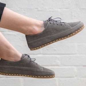 Può includere: Un paio di scarpe in pelle scamosciata grigia con lacci e suola cucita marrone. Le scarpe hanno un aspetto casual e comodo.