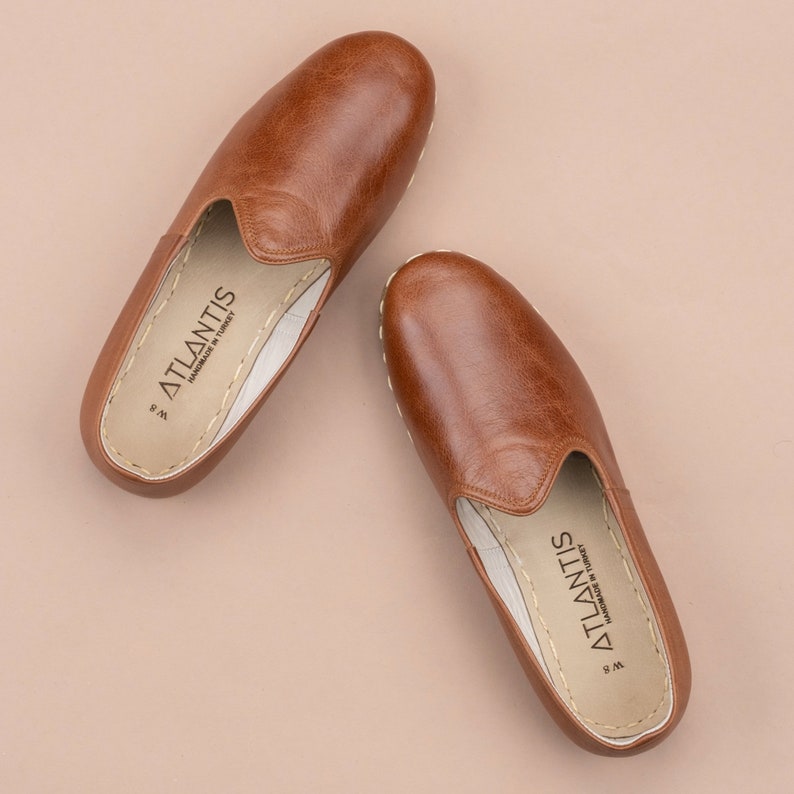 May include: A pair of brown leather slippers with a white sole and the text "ATLANTIS HANDMADE IN TURKEY" on the inside.