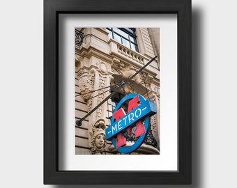 Könnte beinhalten: Ein blaues und rotes "Metro"-Schild hängt an einem Gebäude mit verzierten Details. Das Schild ist ein Kreis mit einem stilisierten "M" darin.