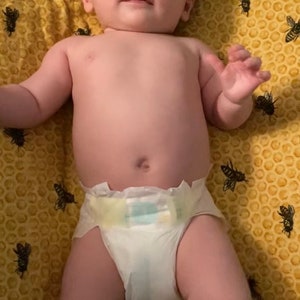 Puede incluir: Un beb&eacute; con un pa&ntilde;al blanco yace sobre una manta amarilla con un patr&oacute;n de panal y abejas negras impresas en ella.