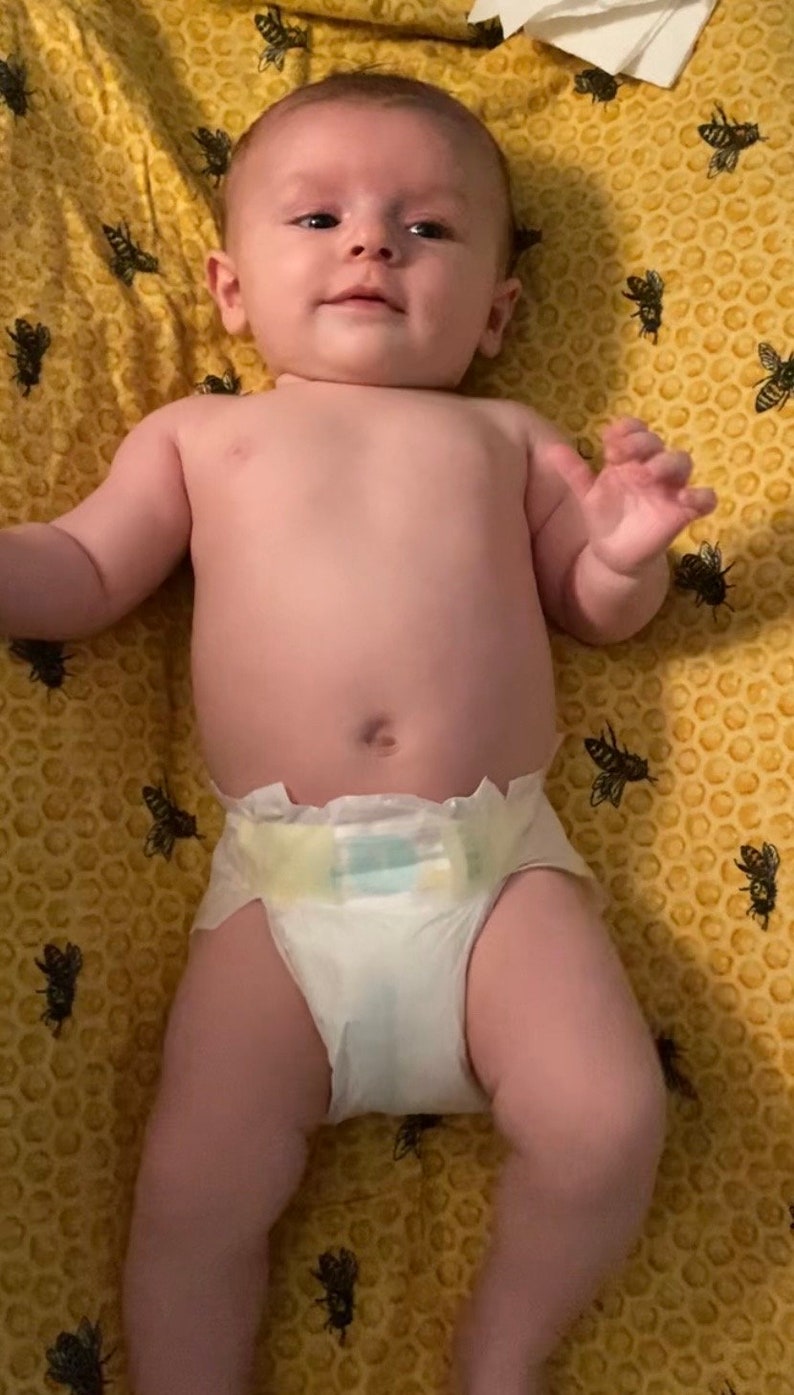 Puede incluir: Un beb&eacute; con un pa&ntilde;al blanco yace sobre una manta amarilla con un patr&oacute;n de panal y abejas negras impresas en ella.