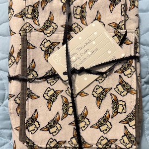 Puede incluir: Una manta de beb&eacute; blanca con un patr&oacute;n repetido de un personaje alien&iacute;gena verde. La manta est&aacute; doblada y atada con una cinta negra. Una etiqueta blanca est&aacute; unida a la cinta con el texto "Baby Blanket, Burp Cloth, 2 Wash clothes".