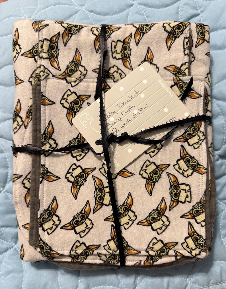 Puede incluir: Una manta de beb&eacute; blanca con un patr&oacute;n repetido de un personaje alien&iacute;gena verde. La manta est&aacute; doblada y atada con una cinta negra. Una etiqueta blanca est&aacute; unida a la cinta con el texto "Baby Blanket, Burp Cloth, 2 Wash clothes".