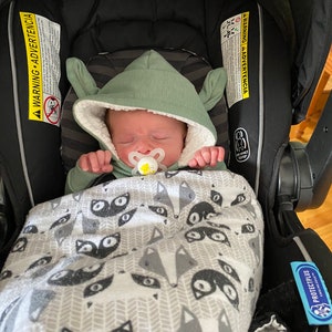 Puede incluir: Un beb&eacute; con un mono verde con capucha y orejeras est&aacute; durmiendo en un asiento de coche negro. El beb&eacute; est&aacute; envuelto en una manta blanca con estampado de zorro negro. El beb&eacute; lleva un chupete.