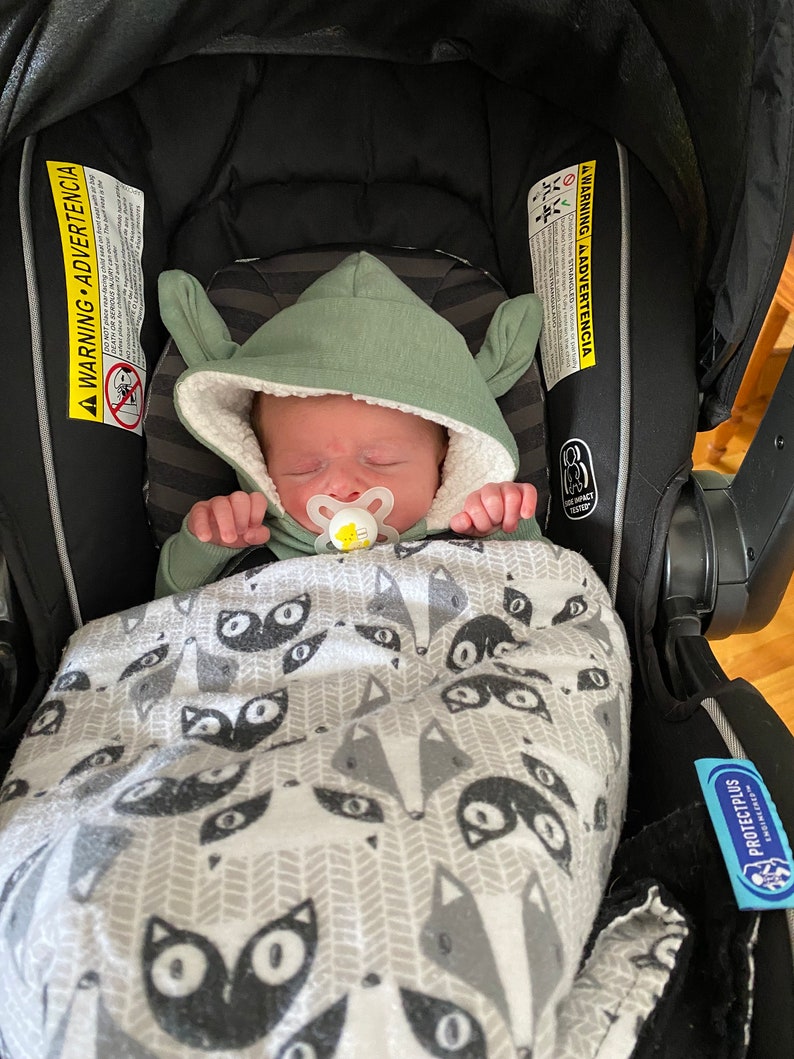 Puede incluir: Un beb&eacute; con un mono verde con capucha y orejeras est&aacute; durmiendo en un asiento de coche negro. El beb&eacute; est&aacute; envuelto en una manta blanca con estampado de zorro negro. El beb&eacute; lleva un chupete.