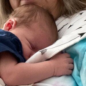 Puede incluir: Una mujer sosteniendo un beb&eacute; dormido en sus brazos. El beb&eacute; lleva una camisa azul y est&aacute; envuelto en una manta blanca con flechas negras impresas. La mujer lleva una camisa azul claro con un patr&oacute;n de flechas blancas y negras.