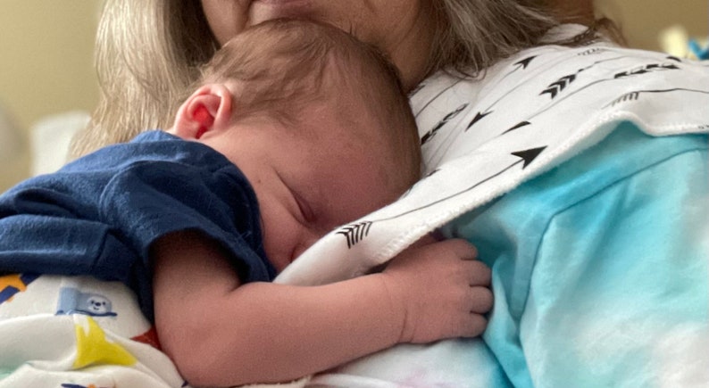 Puede incluir: Una mujer sosteniendo un beb&eacute; dormido en sus brazos. El beb&eacute; lleva una camisa azul y est&aacute; envuelto en una manta blanca con flechas negras impresas. La mujer lleva una camisa azul claro con un patr&oacute;n de flechas blancas y negras.