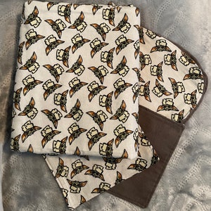 Puede incluir: Una manta de beb&eacute; marr&oacute;n y blanca con un patr&oacute;n repetido de un personaje de dibujos animados con ojos y orejas grandes. La manta est&aacute; hecha de tela suave y tiene un forro de vell&oacute;n marr&oacute;n.