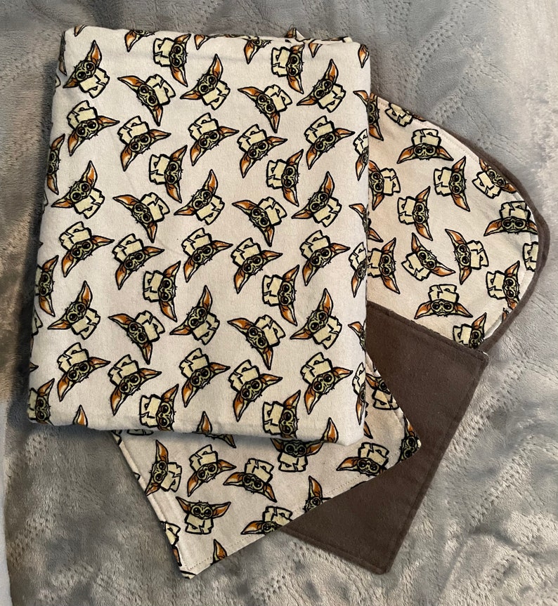 Puede incluir: Una manta de beb&eacute; marr&oacute;n y blanca con un patr&oacute;n repetido de un personaje de dibujos animados con ojos y orejas grandes. La manta est&aacute; hecha de tela suave y tiene un forro de vell&oacute;n marr&oacute;n.