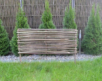 May include: A brown wooden fence made of woven branches, with a similar fence in the background. The fence is in front of a row of evergreen trees.