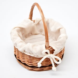 May include: A round wicker basket with a fabric lining and a woven handle. The basket has a white fabric lining with a subtle striped pattern. The handle is made of woven wicker and is attached to the basket with a white fabric tie.