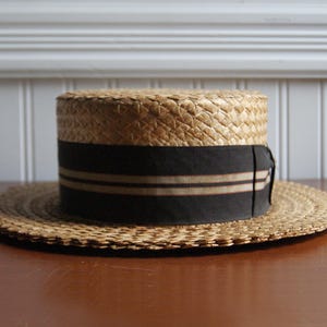 May include: A straw boater hat with a wide, flat brim and a cylindrical crown. The hat features a black band with two thin tan stripes. The hat is made of woven straw and sits on a wooden surface.