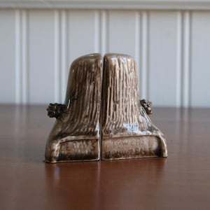 May include: A pair of ceramic bookends, each in the shape of a tree trunk. The bookends are brown with a textured surface, and a small decorative element is attached to the side. The base of each bookend has text.
