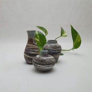 May include: Three ceramic vases in different sizes and shapes, with a marbled brown, white, and grey design. The vases have a textured appearance and are arranged with green leafy plants in the background. The vases have a blue rim.