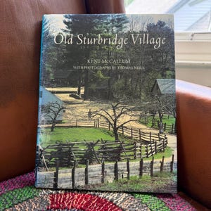 May include: A hardcover book titled Old Sturbridge Village by Kent McCallum with photographs by Thomas Neill. The cover features a color photograph of a rural landscape with wooden split-rail fences, green grass, trees, and historic buildings.