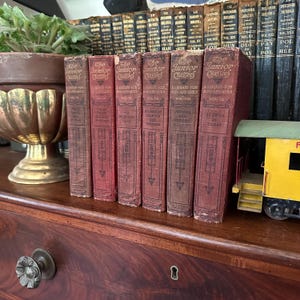 May include: A set of six red hardcover books titled "The Junior Classics" with gold lettering. Each book has a different title and volume number. The books are arranged in a row on a wooden shelf.