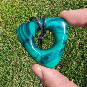 Può includere: Un ciondolo in pietra di malachite verde e nera con un foro al centro, appeso a un cordoncino nero.