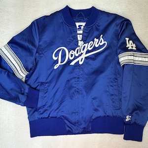 Puede incluir: Una chaqueta bomber de satén azul real con el texto Dodgers en blanco sobre el pecho. La prenda incluye un logotipo LA blanco en la manga izquierda, detalles de rayas blancas y grises en las mangas, cuello, puños y dobladillo de punto acanalado.