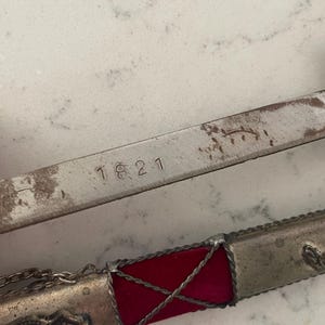 Könnte beinhalten: Nahaufnahme einer antiken Schwertklinge mit der eingravierten Zahl "1821". Der Griff hat einen roten Samtabschnitt, silberne Metallakzente und einen roten Edelstein. Eine kleine Kette ist am Griff befestigt.