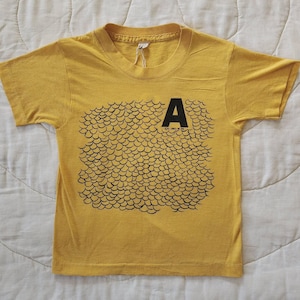 May include: A mustard yellow short-sleeve t-shirt featuring a black graphic print of a repeating scale pattern. The letter A is printed in black at the top right of the scale design. A small white tag is attached to the inside of the crew neckline.