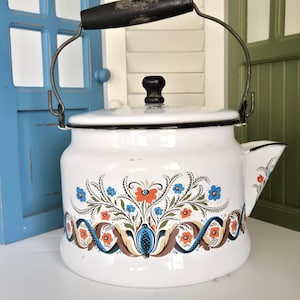 May include: White enamelware kettle with a black handle and lid knob. The kettle features a floral design in blue, orange, and brown. The spout is on the right side, and the handle is arched. A vintage kitchenware item.