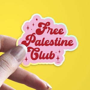 May include: A pink sticker with the words "Free Palestine Club" in a retro font. The sticker has a light green outline and is held against a yellow background.