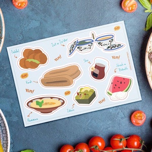 May include: A light blue sticker sheet featuring illustrations of Middle Eastern food items like falafel, hummus, and watermelon. The sheet includes the text "YUM!" and various food names. The background is a blue surface with food items.