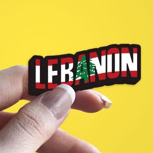 May include: A hand holding a sticker with the word "LEBANON" written in black letters with the Lebanese flag in the background.