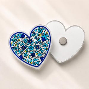 May include: Two heart-shaped acrylic magnets. One magnet features a detailed floral pattern in shades of light blue, dark blue, and yellow on a white background. The other magnet is plain white with a circular metallic magnet on the back.