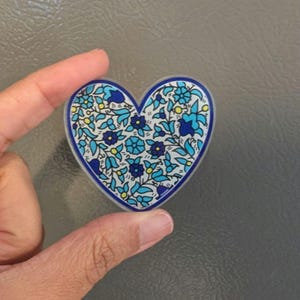 May include: Heart-shaped magnet with a floral design. The magnet features a white background with blue and light blue flowers and leaves, outlined with a dark blue border. Yellow accents are scattered throughout the design.
