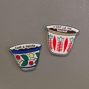 Op de afbeelding: Twee decoratieve koffiekopmagneten. Eén kopje is wit met rode en groene ontwerpen en de tekst "C'EST LA VIE HABIBI". Het andere kopje is blauw met kleurrijke bloemmotieven en Arabische tekst.
