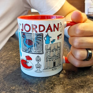 Könnte beinhalten: Weiße Keramik-Kaffeetasse mit rotem Rand und Henkel. Die Tasse zeigt eine farbenfrohe Illustration verschiedener Sehenswürdigkeiten und Symbole aus Jordanien, darunter der Text "JORDAN", "MADABA" und "PETRA".