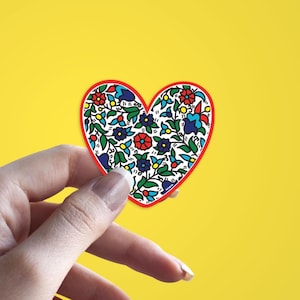May include: A heart-shaped sticker with a red border and a floral pattern. The design features red, blue, and yellow flowers with green leaves on a white background. The sticker is held against a yellow background.