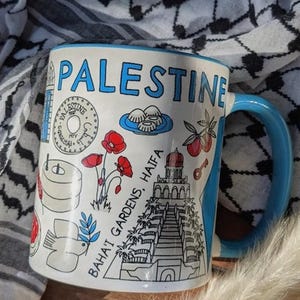 Könnte beinhalten: Weißer Keramikbecher mit blauem Henkel und Rand, mit dem Wort "PALESTINE" in Blau. Der Becher ist mit Illustrationen lokaler Sehenswürdigkeiten, Essen und Blumen verziert. Der Hintergrund umfasst einen schwarz-weiß gemusterten Schal.