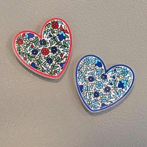 May include: Two heart-shaped magnets with floral patterns. One has a red border and features red, blue, and green flowers on a white background. The other has a blue border and blue flowers.