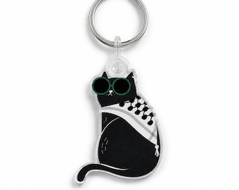 May include: A keychain featuring a black cat illustration wearing round green sunglasses and a white and black patterned keffiyeh scarf. The cat is depicted in a sitting position with its tail curved, attached to a circular silver metal ring.