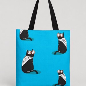 May include: A bright blue tote bag featuring a repeating pattern of black cats wearing red-rimmed sunglasses and white and black patterned scarves. The bag has two black fabric shoulder straps attached to the top edge for carrying items.