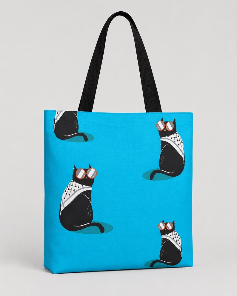 May include: A bright blue tote bag featuring a repeating pattern of black cats wearing red-rimmed sunglasses and white and black patterned scarves. The bag has two black fabric shoulder straps attached to the top edge for carrying items.